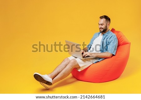 Full body young smiling happy man 20s wear blue shirt white t-shirt sit in bag chair hold use work on laptop pc computer isolated on plain yellow background studio portrait. People lifestyle concept.