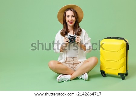 Full length fun happy traveler tourist woman in casual clothes hat suitcase sit taking picture by camera isolated on green background Passenger travel abroad weekend getaway Air flight journey concept