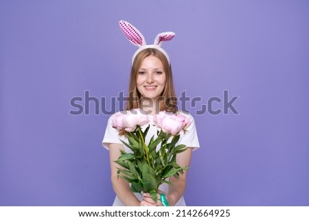 Charming happy cute girl with pink easter bunny ears and bouquet spring peonies, in white t-shirt posing on purple background up to her waist. Woman smiles and rejoices at arrival spring and easter
