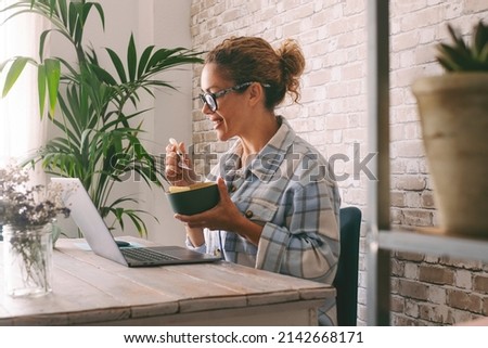 Side view of adult young woman smiling and eating in fron tof a laptop at the desk at home. Alternative office lifestyle female people working in video call conference during lunch time. Modern job Royalty-Free Stock Photo #2142668171
