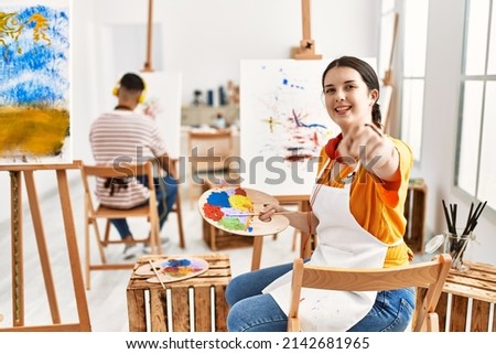 Young artist woman painting on canvas at art studio pointing to you and the camera with fingers, smiling positive and cheerful 