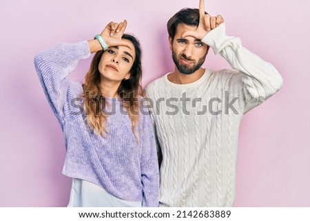 Young hispanic couple wearing casual clothes making fun of people with fingers on forehead doing loser gesture mocking and insulting. 