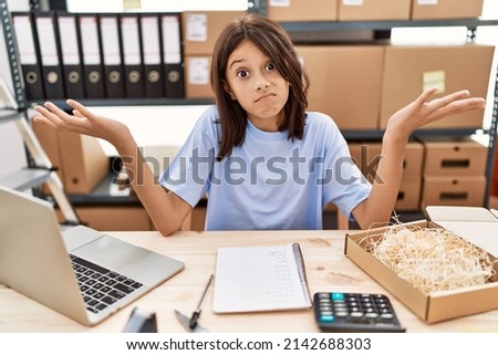Young hispanic girl working at small business ecommerce clueless and confused expression with arms and hands raised. doubt concept. 