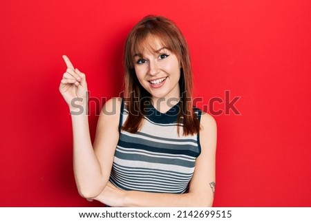 Redhead young woman wearing casual t shirt with a big smile on face, pointing with hand and finger to the side looking at the camera. 