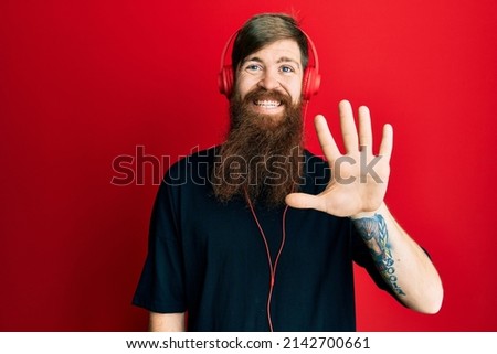 Redhead man with long beard listening to music using headphones showing and pointing up with fingers number five while smiling confident and happy. 