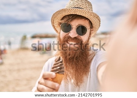 Young redhead tourist man eating ice cream making selfie by the camera at the beach.