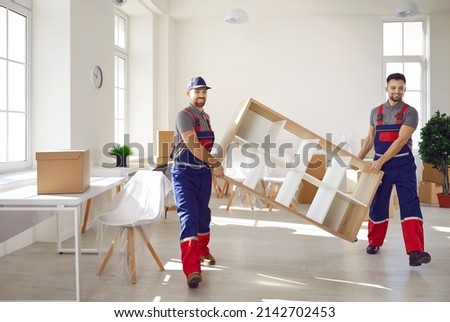Truck delivery service workers in workwear uniforms removing furniture from house or apartment. Two happy young men from moving company carrying bookshelf together Royalty-Free Stock Photo #2142702453