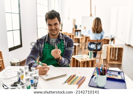 Young artist man at art studio beckoning come here gesture with hand inviting welcoming happy and smiling 