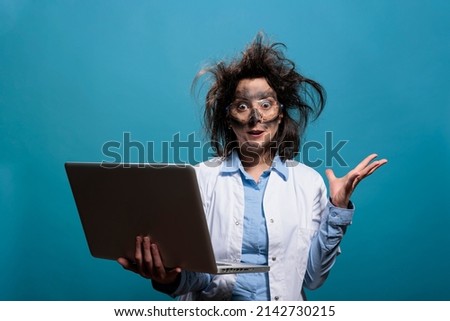 Lunatic chemist with messy hair and dirty face having laptop while gesticulating angrily after dangerous lab explosion. Insane scientist being enraged after failed laboratory chemical experiment. Royalty-Free Stock Photo #2142730215