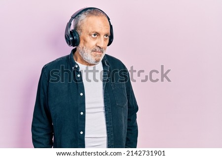 Handsome senior man with beard listening to music using headphones smiling looking to the side and staring away thinking. 