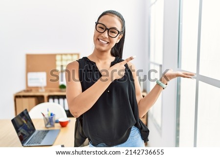 Young hispanic business woman working at the office amazed and smiling to the camera while presenting with hand and pointing with finger. 