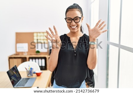 Young hispanic business woman working at the office showing and pointing up with fingers number nine while smiling confident and happy. 