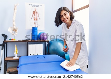Young latin woman wearing physiotherapist uniform cleaning massage table at physiotherapy clinic