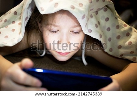 Cute little girl hiding under a blanket and watching a cartoon about magical space on the phone.
