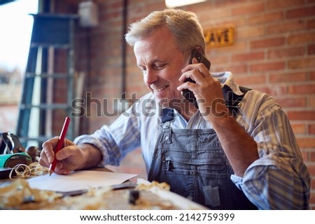 Mature Male Carpenter In Garage Workshop Working On Design And Talking On Mobile Phone