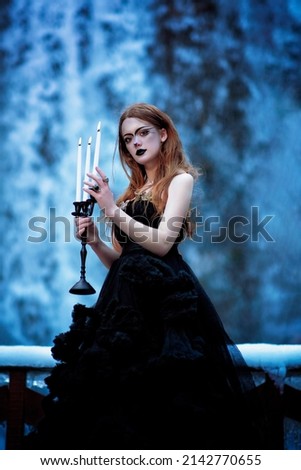 A girl with red hair stands against the backdrop of a winter blue waterfall. A strange girl with an unusual make-up. A gothic woman in a black dress with a candlestick in her hands and burning candles