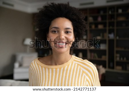 Happy pretty young African American woman in casual home head shot portrait. Millennial curly haired girl with toothy smile, curly hair posing in apartment interior, laughing, looking at camera