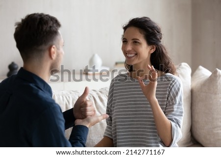 Smiling millennial Latina woman communicates with man use sign language sit together on sofa at home. Couple or friends with disability talking, showing fingers gestures share news, enjoy conversation