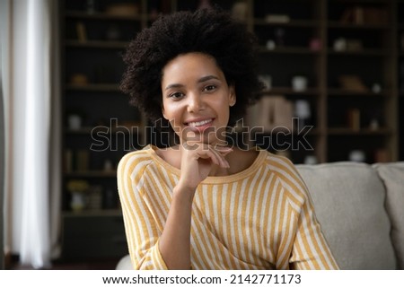 Happy millennial African American girl home head shot portrait. Smiling young woman looking at camera, talking on video call, sitting on couch, speaking online, touching face. Screen view
