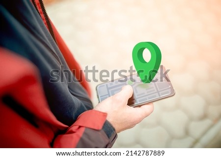 Online navigation, GPS and Geocaching. Green geolocation sign above the smartphone screen with an abstract online map. Close-up of a man's hand using a smartphone on the street. Copy space Royalty-Free Stock Photo #2142787889