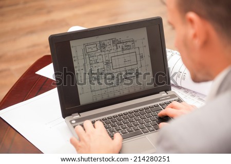Close-up portrait of laptop with blueprints, architect sitting from behind working on architectural plan, interior shot Royalty-Free Stock Photo #214280251