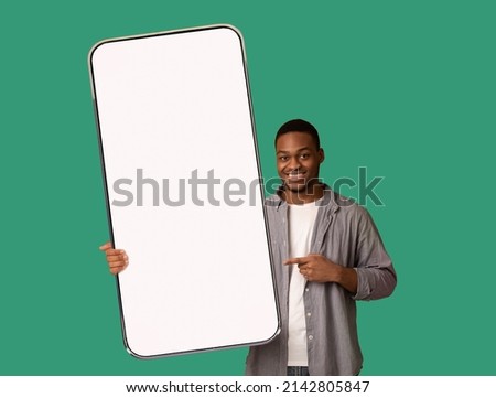 Great mobile offer. Happy black man pointing at huge empty smartphone in his hand, advertising new app or website, showing mockup for your design, standing over green background