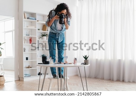 Photography And Makeup Blogging. Professional Female Photographer Using Camera Taking Photos Of Beauty Products Standing On Chair Near Desk Working In Photo Studio. Full Length Shot