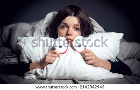 Girl hugs a pillow with tired red eyes and a sad expression on her face, hides under the covers. Woman is worried about insomnia, poor sleep, stress, in her hands a creative pillow with paper eyes.