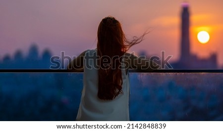Woman looking and enjoying the sunset view from balcony with the sun setting behind skyscraper in busy urban downtown with loneliness for solitude, loneliness and dreaming of freedom lifestyle concept Royalty-Free Stock Photo #2142848839