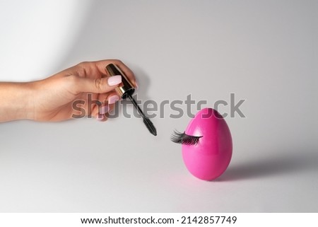Woman’s hand holding eye mascara to apply on lashes on pink Easter egg. Make up and beauty Easter concept. Creative minimal Easter composition.                                Royalty-Free Stock Photo #2142857749