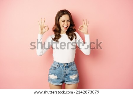 Stylish glamour girl with dark curly hairstyle, showing okay gesture, ok satisfied sign, smiling pleased, complimenting, recommending something good, pink background