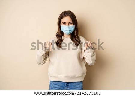 Cheerful cute woman in medical face mask, pointing fingers down, showing advertisement, using protection from covid-19, beige background