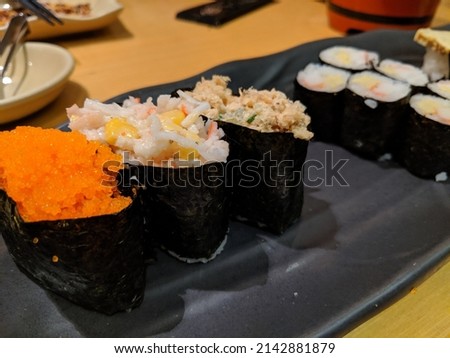 Black tray with some pieces of sushi accompanied by salads and vegetables. Next door we have some dishes. Items are on a brown table. In the picture we have the colors white, red . 