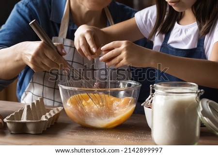 Grandkid helping grandma to cook omelet for breakfast, cracking beating eggs with whisker in bowl at kitchen table. Grandmother teaching grandchild to bake, preparing dough. Family activity. Close up Royalty-Free Stock Photo #2142899997