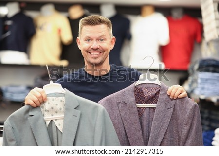 Smiling man is holding two suits on hangers in store. Choosing business men suit concept