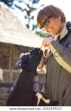 Happy smiling woman feeding black goat with carrots, special food, treats for animals in nursery, contact zoo, eco park. Animal care on a farm in a village. Human and nature interaction vertical photo