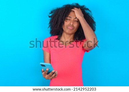 Upset depressed Young woman with afro hairstyle wearing pink t-shirt over blue background makes face palm as forgot about something important holds mobile phone expresses sorrow and regret blames