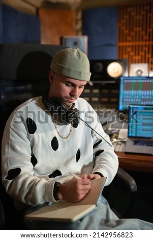 Portrait of confident young music producer writing in notepad. Man wearing cap and stylish white sweatshirt working in sound recording studio. Creating music concept