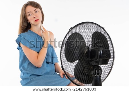 Woman is sitting in front of fan. She wipes off sweat napkin. Concept is torment from heat. Girl with fan on white background. Woman in denim dress. She cools herself electric fan.