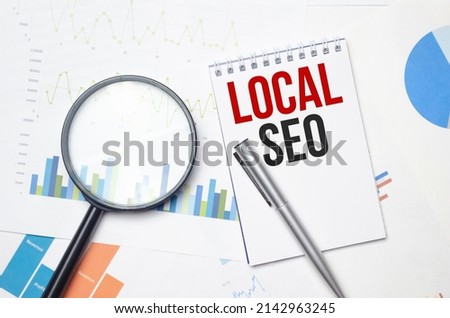 Magnifying glass loupe with sign LOCAL SEO between two paper sheets on the blue table.
