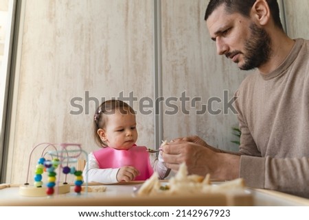 A father is feeding his daughter  inside the house during the day