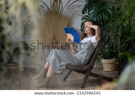 Young woman waving with paper fan while sitting in home garden filled with tropical foliage plants, overheated female fanning herself to cool down escaping heat in greenhouse on hot summer day Royalty-Free Stock Photo #2142986545