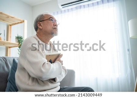 Asian senior male sitting alone on sofa and looks family photographs. Elderly older mature grandfather feel lonely and sad, missing his relative and wife while stay in nursing home after retirement. Royalty-Free Stock Photo #2143009449