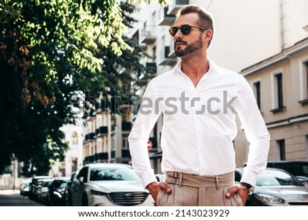 Portrait of handsome confident stylish hipster lambersexual model.Sexy modern man dressed in white shirt and trousers. Fashion male posing on street background in Europe city at sunset. In sunglasses Royalty-Free Stock Photo #2143023929