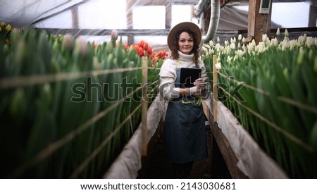 Portrait of beautiful young female smiling woman worker florist in apron stands with tablet in greenhouse and looks at camera