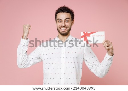 Smiling joyful young bearded man 20s wearing basic casual white shirt standing doing winner gesture hold gift certificate looking camera isolated on pastel pink color wall background studio portrait