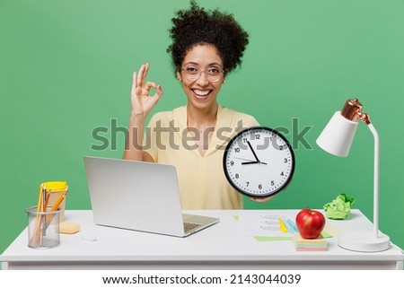 Young employee business woman of African American ethnicity in shirt sit work at white office desk with pc laptop hold in hands clock show ok isolated on plain green background. Achievement concept