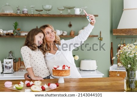 Happy chef cook baker mom woman in white shirt work with baby girl helper do selfie shot on mobile cell phone at kitchen table home Cooking food process concept Mommy little kid daughter prepare cake.