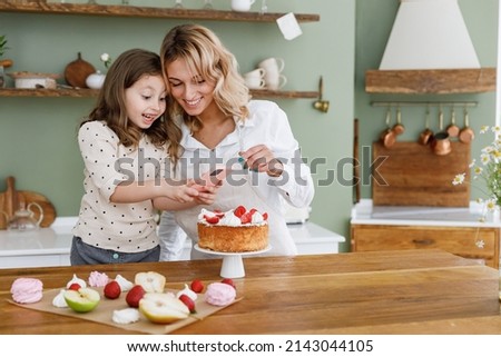 Chef cook baker mom woman in white shirt work with amazed baby girl helper take picture on mobile cell phone at kitchen table home. Cooking food process concept Mommy little kid daughter prepare cake.