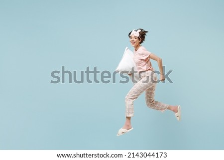 Full length side view young happy excited woman in pajamas jam sleep eye mask rest relaxing at home jump high with pillow run isolated on pastel blue background studio. Good mood night bedtime concept Royalty-Free Stock Photo #2143044173